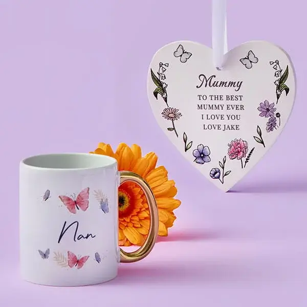 White butterfly‑themed Nan mug displayed beside an orange flower and a hanging heart‑shaped plaque decorated with floral illustrations and a loving message.